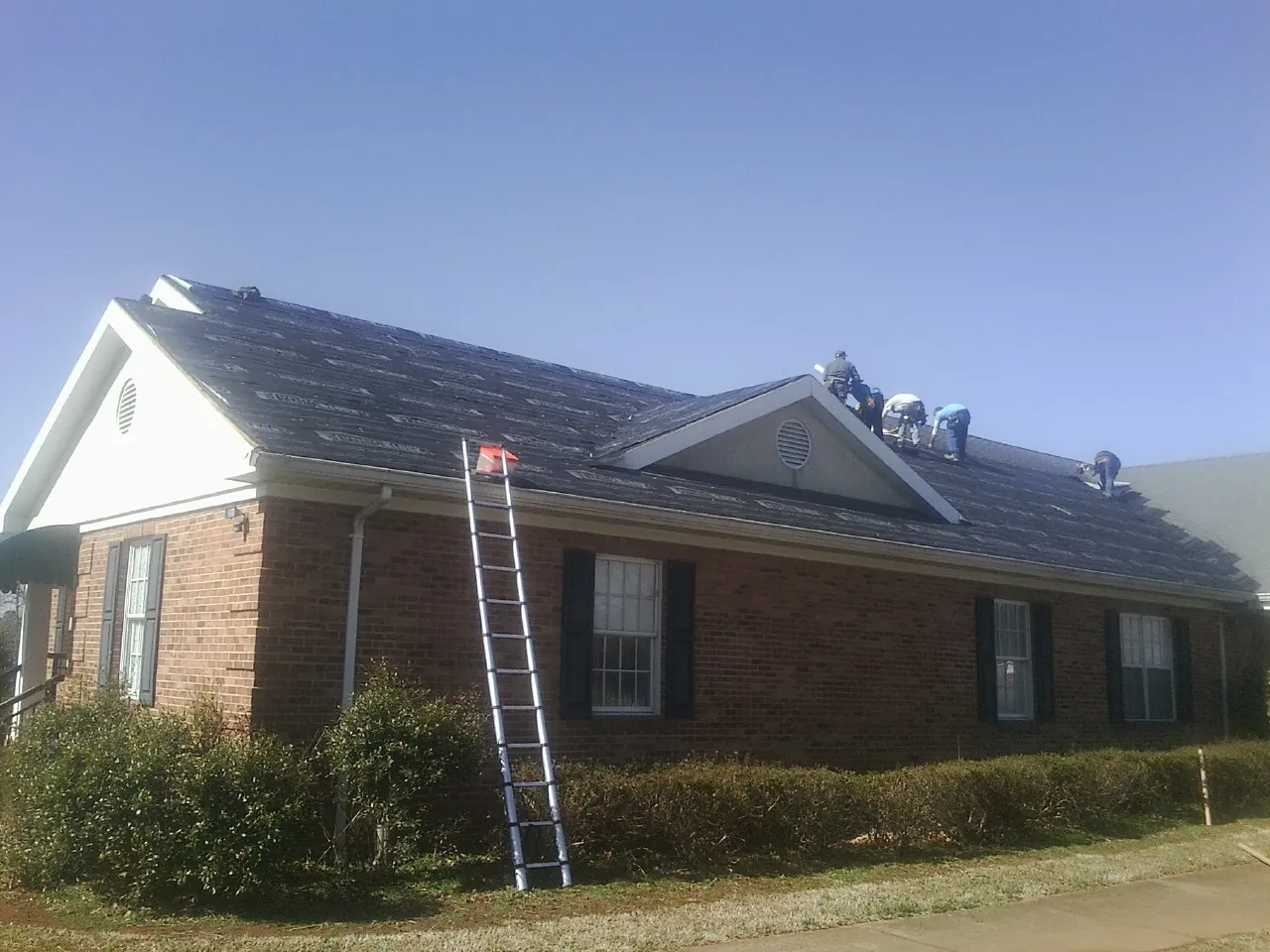 Crew tearing shingles off a brick ranch roof with a ladder set — Metal Roof Repair work by our crew in West Newton